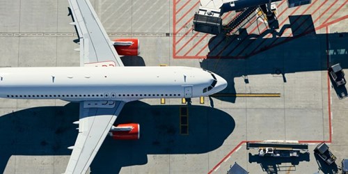 Aerial view of a commercial airplane parked on an airport tarmac. The aircraft is white with red engine covers and is positioned parallel to the ground markings. Surrounding the plane are service vehicles and equipment, including a mobile stairway and baggage carts. The concrete surface features red and yellow safety lines and designated zones. Strong shadows from the plane and equipment are cast across the ground, indicating bright sunlight.