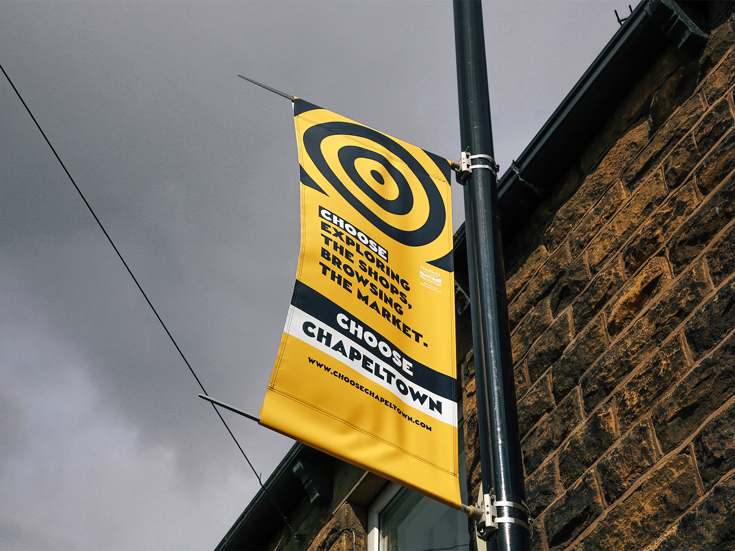 Yellow and black banner attached to a lamppost on a stone building, featuring concentric circle design and text that reads: “Choose exploring the shops, browsing the market. Choose Chapeltown” with a website link www.choosechapeltown.com. Overhead wires and a cloudy sky are visible in the background.