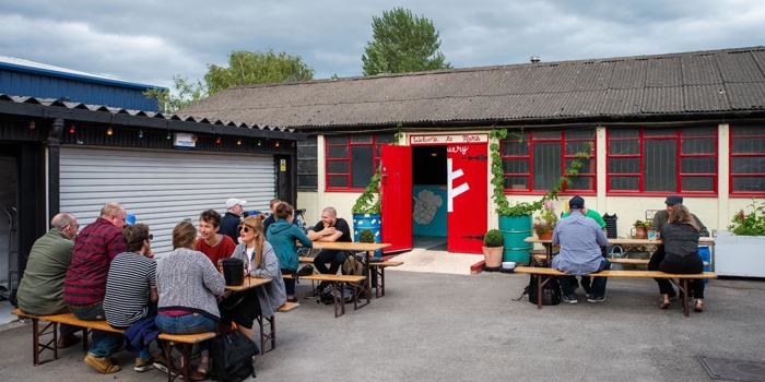 Outdoor seating area with wooden picnic tables and benches, occupied by groups of people. A rustic building with red-framed windows and a red door is in the background, with potted plants near the entrance under an overcast sky.