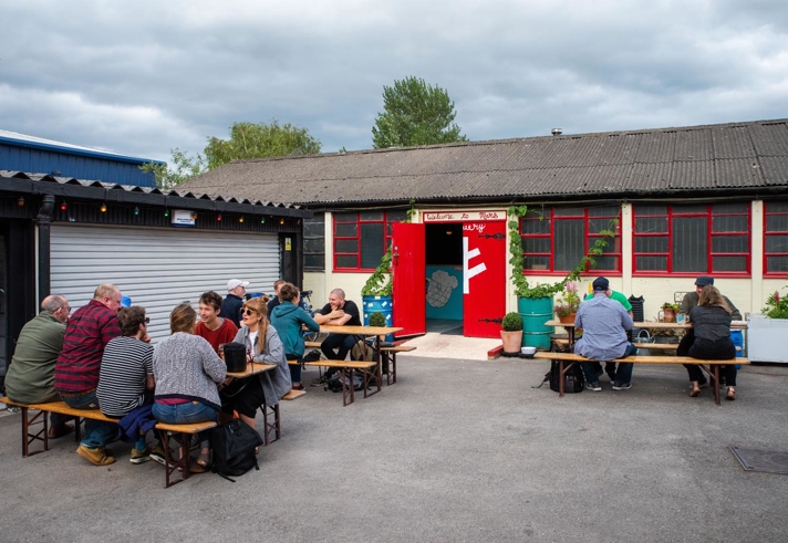 Outdoor seating area with wooden picnic tables and benches, occupied by groups of people. A rustic building with red-framed windows and a red door is in the background, with potted plants near the entrance under an overcast sky.