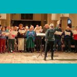 A group of people stand in a row inside a large hall with tall stone columns, each holding sheet music while participating in a choral rehearsal. A person stands in front of the group with raised arms, appearing to lead the singers, while another person sits to the right at a keyboard instrument. The scene is warmly lit, with chairs, music stands, and architectural details of the hall visible in the background, all set within a turquoise border.