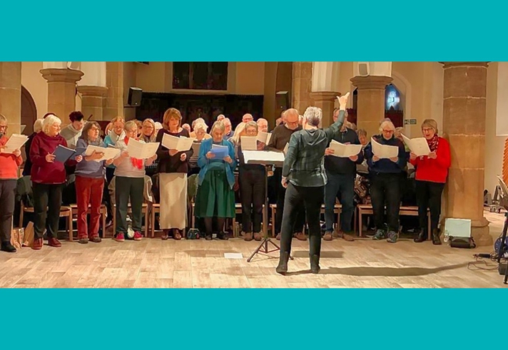 A group of people stand in a row inside a large hall with tall stone columns, each holding sheet music while participating in a choral rehearsal. A person stands in front of the group with raised arms, appearing to lead the singers, while another person sits to the right at a keyboard instrument. The scene is warmly lit, with chairs, music stands, and architectural details of the hall visible in the background, all set within a turquoise border.