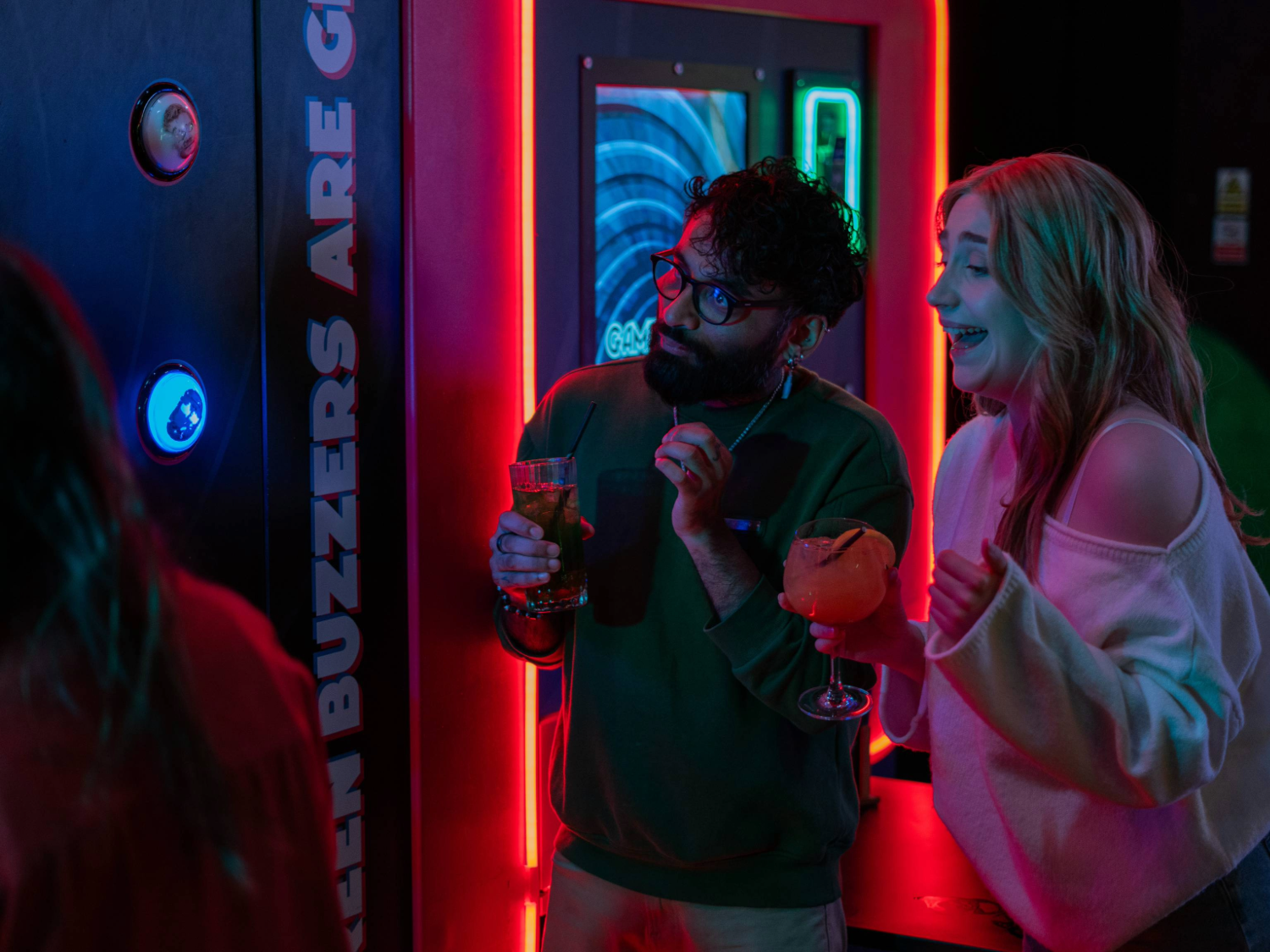 Two people standing near a neon-lit arcade game with glowing buttons and a bright display. Both are holding drinks in cocktail glasses, one orange and one clear, while watching the game. The machine features colourful lights and text reading “Green buzzers are great.” The setting is vibrant and modern, with a lively atmosphere and illuminated signage in the background.