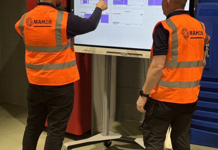Two people wearing bright orange safety vests with the Maher logo stand in front of a large interactive screen in an industrial setting. One person is pointing at charts displayed on the screen, while the other observes. The background includes a red cabinet and blue storage units.