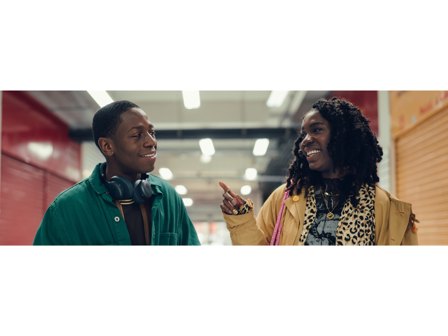 Two people standing in an indoor setting with bright overhead lights and red and orange walls. One person wears a green jacket over a dark top with headphones around the neck. The other person wears a tan coat layered over a leopard-print scarf and graphic T-shirt, accessorised with gold jewellery and a pink strap bag. The second person gestures with one hand while speaking, suggesting a casual conversation in a corridor-like space.