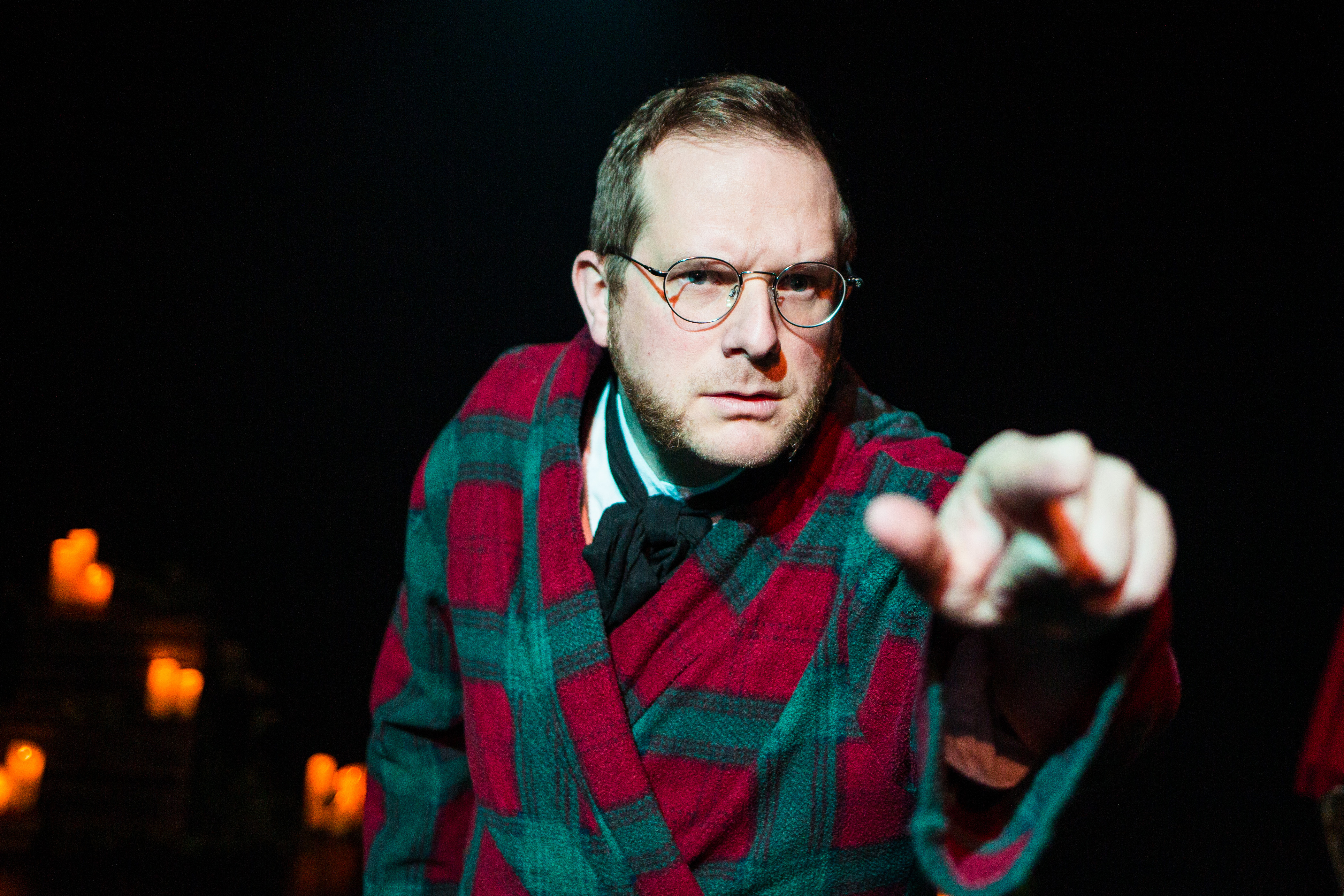 Person wearing a red and green checked robe over a white shirt and dark scarf, standing in a dimly lit setting and pointing towards the camera. Warm candlelight glows in the background, creating a dramatic atmosphere against the dark backdrop.