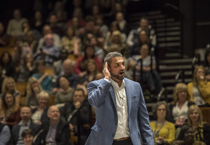 Person in a blue blazer standing in front of a large seated audience in an auditorium. The individual is holding one hand near the ear, possibly listening or engaging with the crowd. The background shows rows of people seated on tiered benches, with steps and railings visible on the right side. Lighting is focused on the person in the foreground, creating a contrast with the dimmer audience area.
