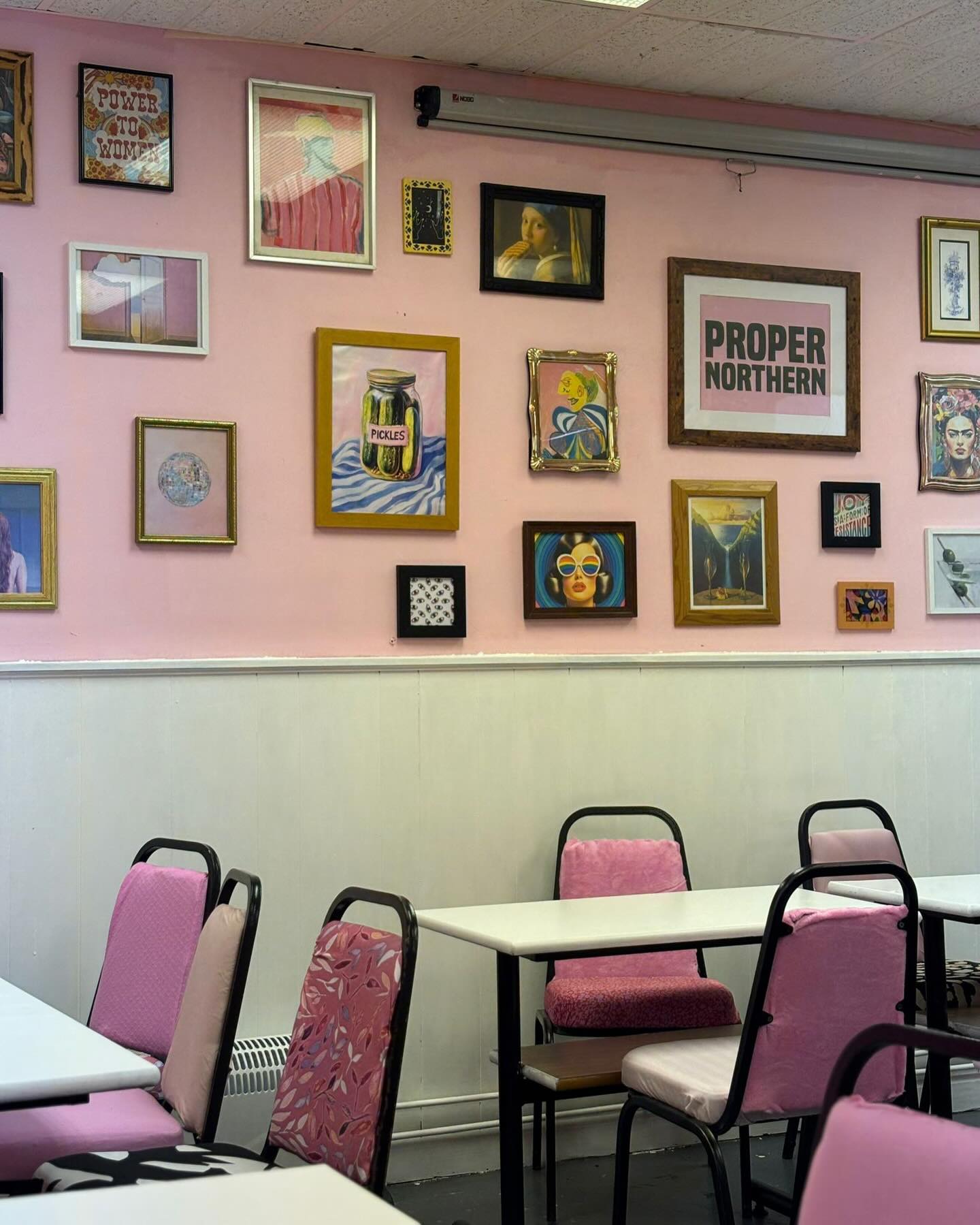 A pastel pink café-style room with white tables and pink chairs, featuring a gallery wall of eclectic framed artwork and a large “Proper Northern” print.