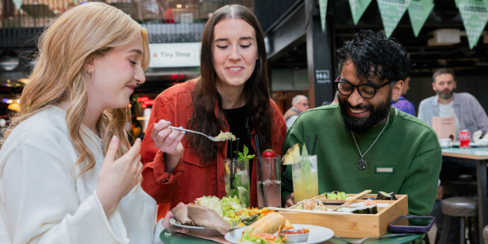 Three people seated at a round green table in an indoor food hall, sharing a meal. The table holds plates of tacos, salad, bread, and a wooden tray with assorted dishes. There are also tall glasses of colourful drinks garnished with fruit and herbs. In the background, green triangular banners hang overhead, and signs for “Oktoberfest” and “Baity” are visible, creating a lively, casual dining atmosphere.