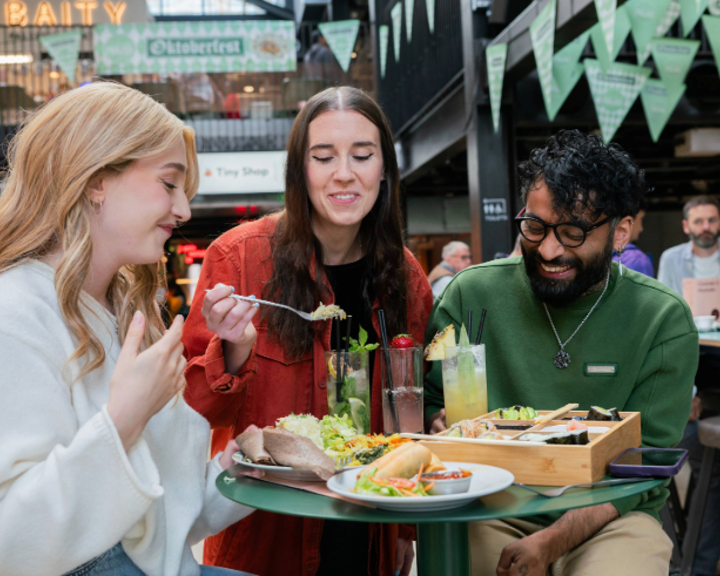 Three people seated at a round green table in an indoor food hall, sharing a meal. The table holds plates of tacos, salad, bread, and a wooden tray with assorted dishes. There are also tall glasses of colourful drinks garnished with fruit and herbs. In the background, green triangular banners hang overhead, and signs for “Oktoberfest” and “Baity” are visible, creating a lively, casual dining atmosphere.
