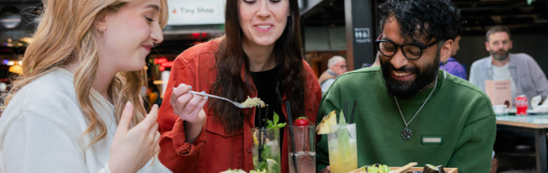 Three people seated at a round green table in an indoor food hall, sharing a meal. The table holds plates of tacos, salad, bread, and a wooden tray with assorted dishes. There are also tall glasses of colourful drinks garnished with fruit and herbs. In the background, green triangular banners hang overhead, and signs for “Oktoberfest” and “Baity” are visible, creating a lively, casual dining atmosphere.