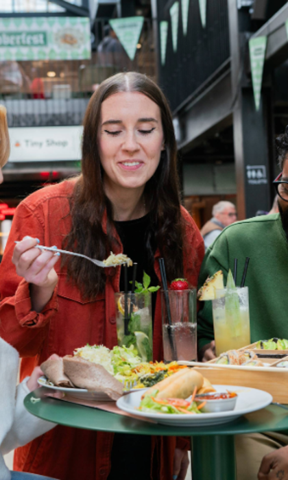 Three people seated at a round green table in an indoor food hall, sharing a meal. The table holds plates of tacos, salad, bread, and a wooden tray with assorted dishes. There are also tall glasses of colourful drinks garnished with fruit and herbs. In the background, green triangular banners hang overhead, and signs for “Oktoberfest” and “Baity” are visible, creating a lively, casual dining atmosphere.