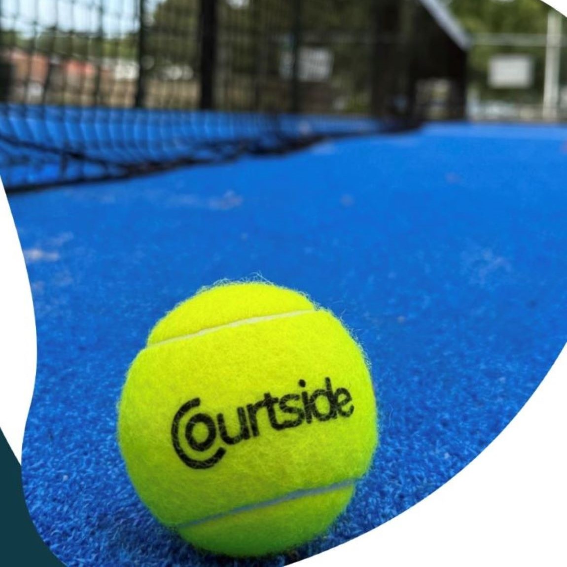 Close-up of a bright yellow tennis ball with the word “Courtside” printed on it, resting on a vibrant blue padel court surface. The black net is visible in the background, slightly out of focus, with outdoor surroundings beyond the court.