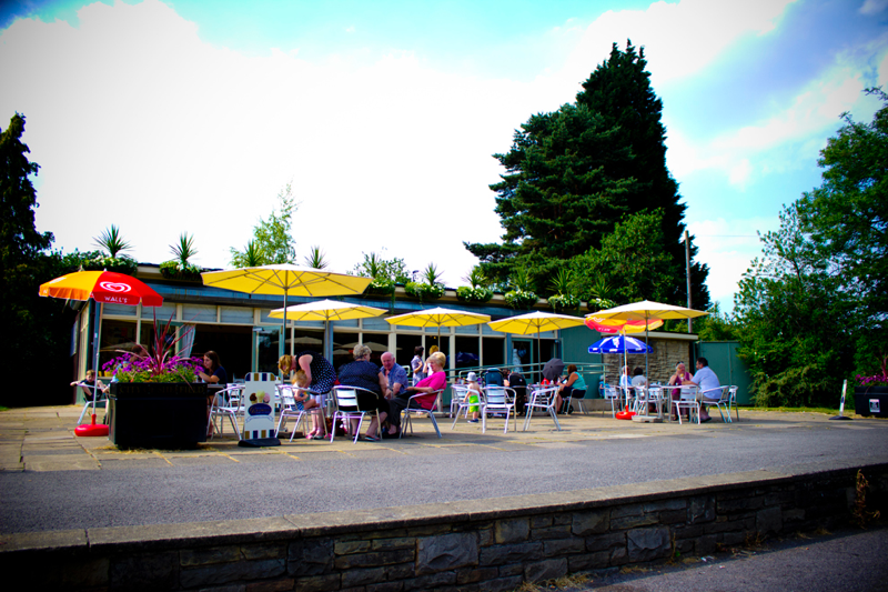 Exterior of Millhouses Park Cafe in the sunshine. There are tables outside with colourful parasols up.
