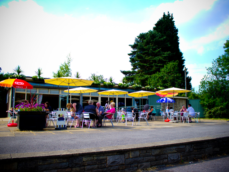 Exterior of Millhouses Park Cafe in the sunshine. There are tables outside with colourful parasols up.