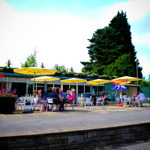 Exterior of Millhouses Park Cafe in the sunshine. There are tables outside with colourful parasols up.
