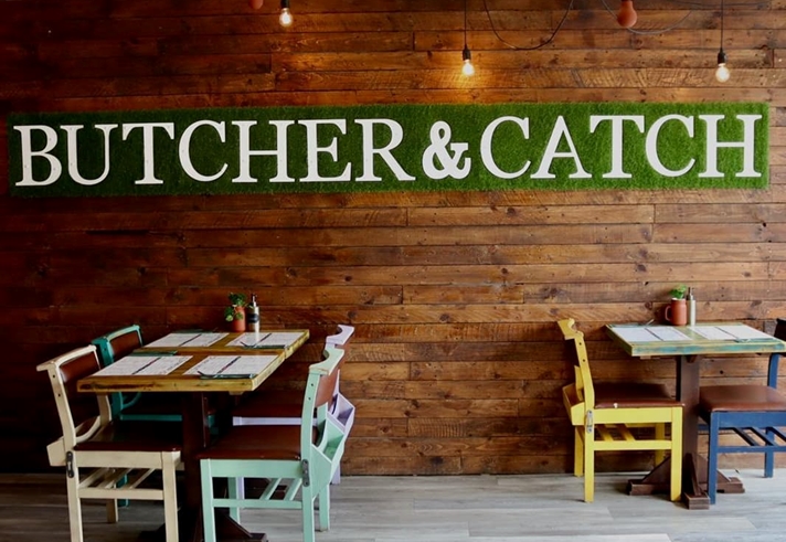 Restaurant tables next to a naked brick wall with a green sign that reads 'Butcher & Catch'.