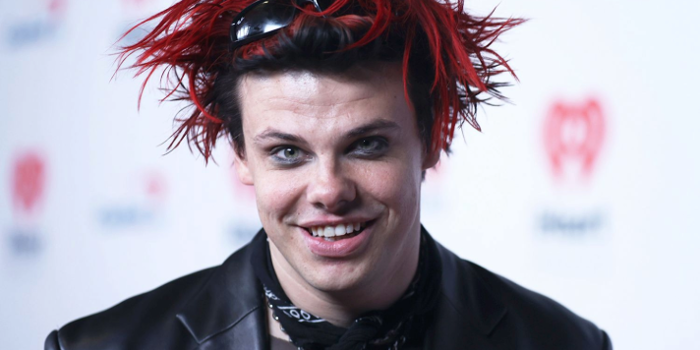 Person wearing a black leather jacket and layered necklaces, including a padlock pendant, with a black scarf tied around the neck. Hair is styled in spiky red and black strands with sunglasses resting on top. The background features a white backdrop with red heart-shaped logos.