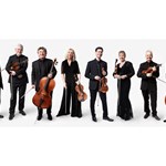 The Academy Of St Martin In The Fields Chamber Ensemble pose against a stark background. They are all wearing black and holding their instruments.