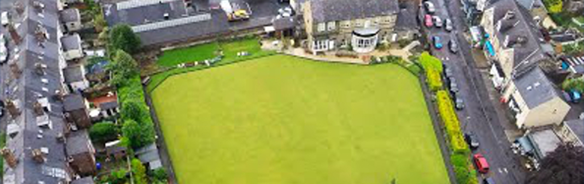 Aerial view of a rectangular green lawn, possibly a bowling green, surrounded by residential houses and narrow streets. The area includes trees, gardens, and parked cars, with a mix of brick buildings and greenery visible throughout the neighbourhood.