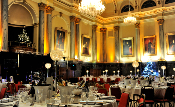 Grand banquet hall with ornate architecture, high arched ceilings, and large chandeliers casting warm light. The room is decorated with framed portraits on the walls and two illuminated Christmas trees. Round tables are elegantly set with white tablecloths, glassware, menus, and champagne bottles in ice buckets. Red chairs surround the tables, and tall crystal centrepieces add a festive touch to the formal setting.