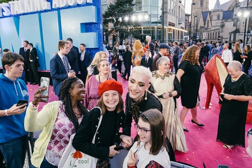 Three people smile and take a selfie together at a red carpet event. Behind them, other attendees in both formal and casual attire mingle. Large blue letters spelling 'ME' are visible, likely part of a sign reading 'EVERYBODY'S TALKING ABOUT JAMIE.' The event takes place outdoors with buildings and lights in the background.
