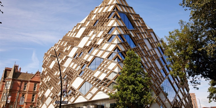 Modern pyramid-shaped building with a striking geometric design featuring glass panels and wooden lattice framework, located on a street corner with trees and older brick buildings nearby under a clear blue sky.