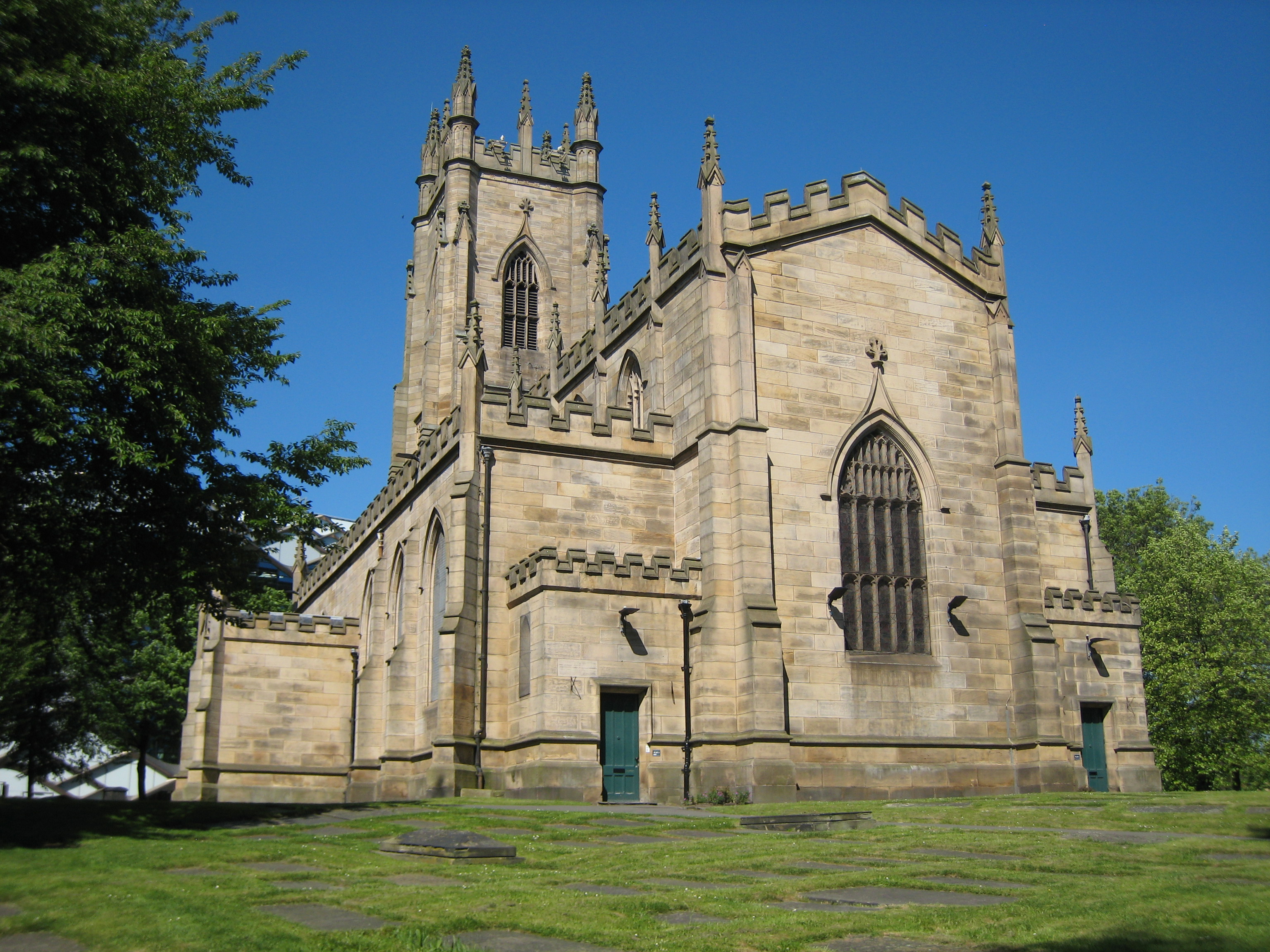 Historic stone church with Gothic architecture, featuring tall arched windows, battlements along the roofline, and a central tower. The building is surrounded by green grass and trees under a clear blue sky.