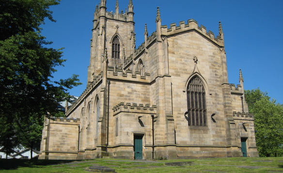 Historic stone church with Gothic architecture, featuring tall arched windows, battlements along the roofline, and a central tower. The building is surrounded by green grass and trees under a clear blue sky.