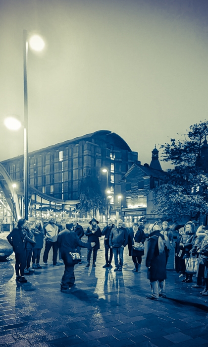 A group of people gathered round a tour guide, in Tudor Square in the centre of Sheffield, at night.