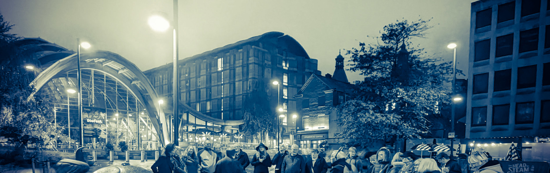 A group of people gathered round a tour guide, in Tudor Square in the centre of Sheffield, at night.
