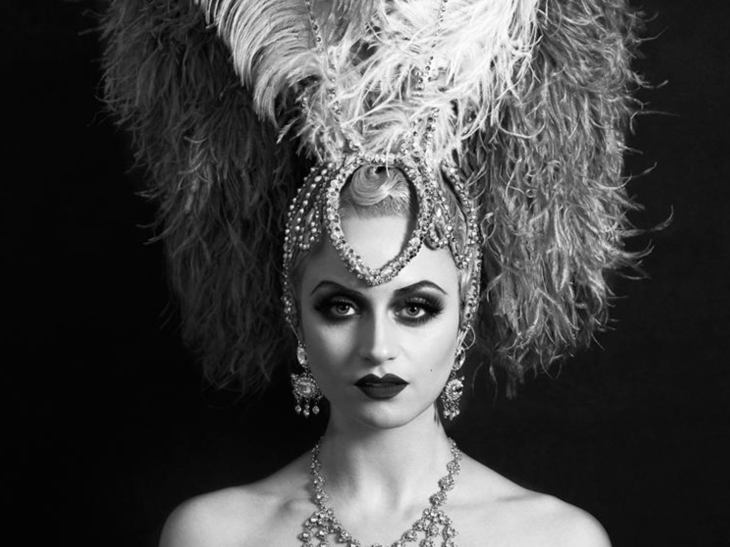 A black‑and‑white portrait of a person wearing an elaborate showgirl-style costume, featuring a large feathered headdress with ornate beading and a tall plume rising from the centre. They also wear a decorative beaded necklace. The background is dark, making the dramatic headdress stand out.