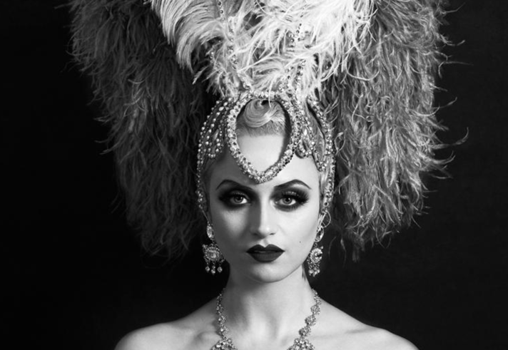A black‑and‑white portrait of a person wearing an elaborate showgirl-style costume, featuring a large feathered headdress with ornate beading and a tall plume rising from the centre. They also wear a decorative beaded necklace. The background is dark, making the dramatic headdress stand out.