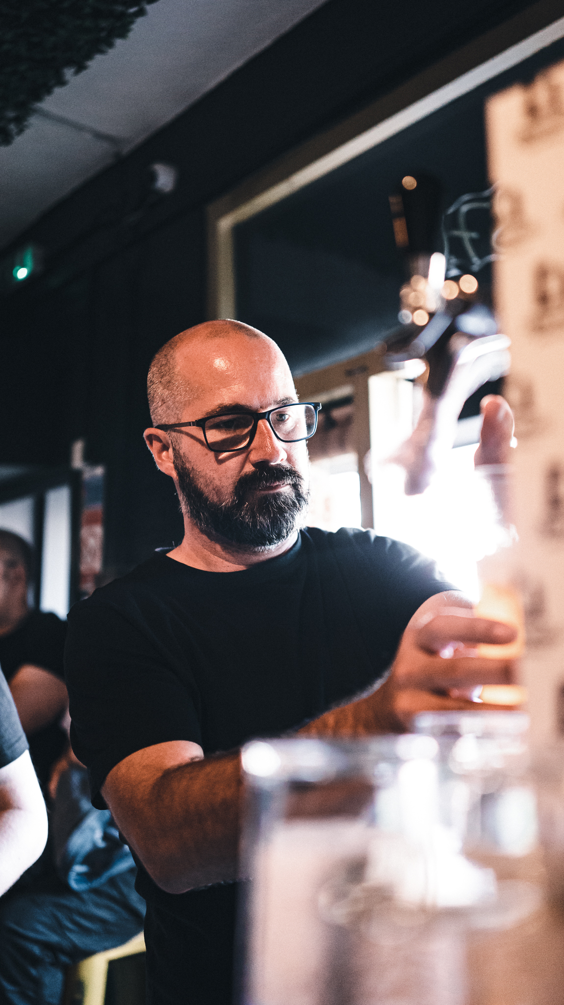 A person pouring a drink at a bar, surrounded by soft warm lighting and bar equipment, with other people visible in the background.
