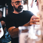 A person pouring a drink at a bar, surrounded by soft warm lighting and bar equipment, with other people visible in the background.