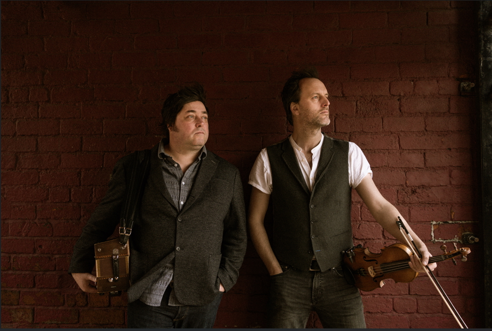 Two musicians stand against a red brick wall, both wearing casual waistcoats and shirts. One holds a violin and bow, while the other carries a small wooden accordion slung over his shoulder. Their relaxed poses suggest a promotional or backstage moment.