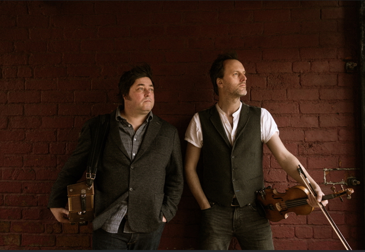 Two musicians stand against a red brick wall, both wearing casual waistcoats and shirts. One holds a violin and bow, while the other carries a small wooden accordion slung over his shoulder. Their relaxed poses suggest a promotional or backstage moment.
