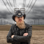 A man in a black leather jackets and a black stetson, stands in the countryside, surrounded by electricity pylons. He has a long beard.
