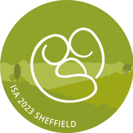 The International Stillbirth Alliance logo - Circular green logo featuring a white abstract line design in the center, with text reading ‘ISA 2023 Sheffield’ along the bottom edge. The background shows a stylized landscape with trees and rolling hills.