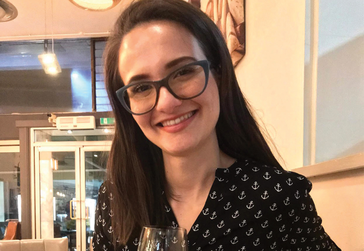 A person seated indoors at a restaurant table, holding a wine glass. The individual is wearing a black shirt patterned with small white anchor designs. The background shows modern décor with wooden panels, ceiling lights, and a glass door leading outside.