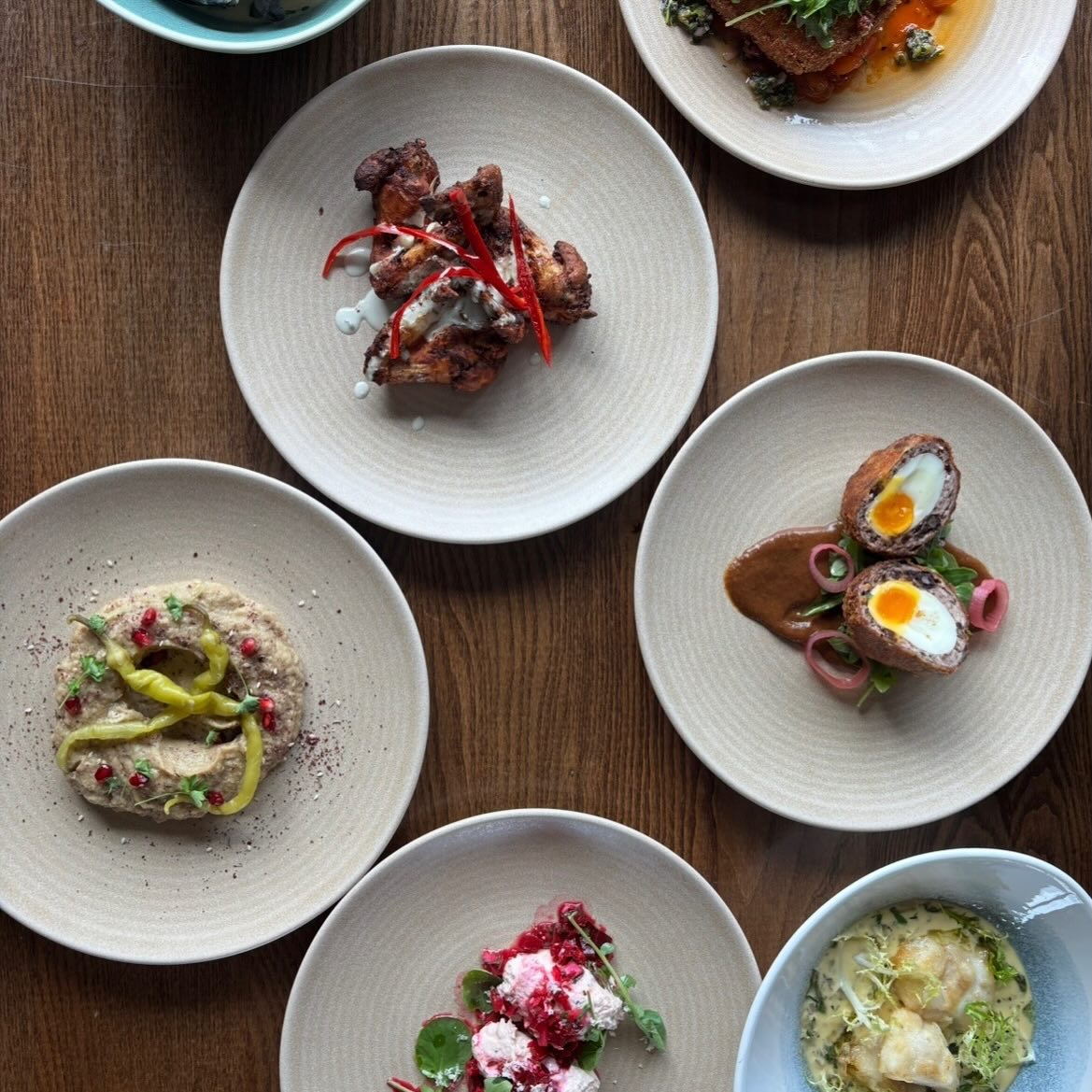 A flat lay of several plated dishes on a wooden table, including a Scotch egg, chicken wings, dips, salads and other small plates.