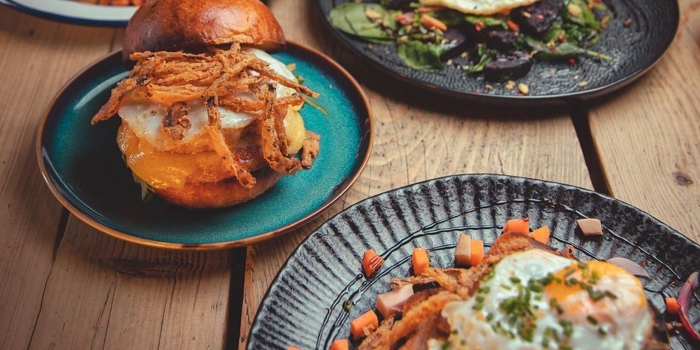 Four plates of breakfast dishes with eggs, sandwiches, greens, and a full English on a wooden table.