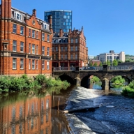 A scenic view of a river flowing through a city with a small weir in the foreground. On the left, there are historic red-brick buildings with ornate architectural details, reflected in the calm water. A stone bridge with three arches spans the river, and behind it rises a modern glass high-rise building against a clear blue sky. Green shrubs line the riverbank, adding a touch of nature to the urban setting.