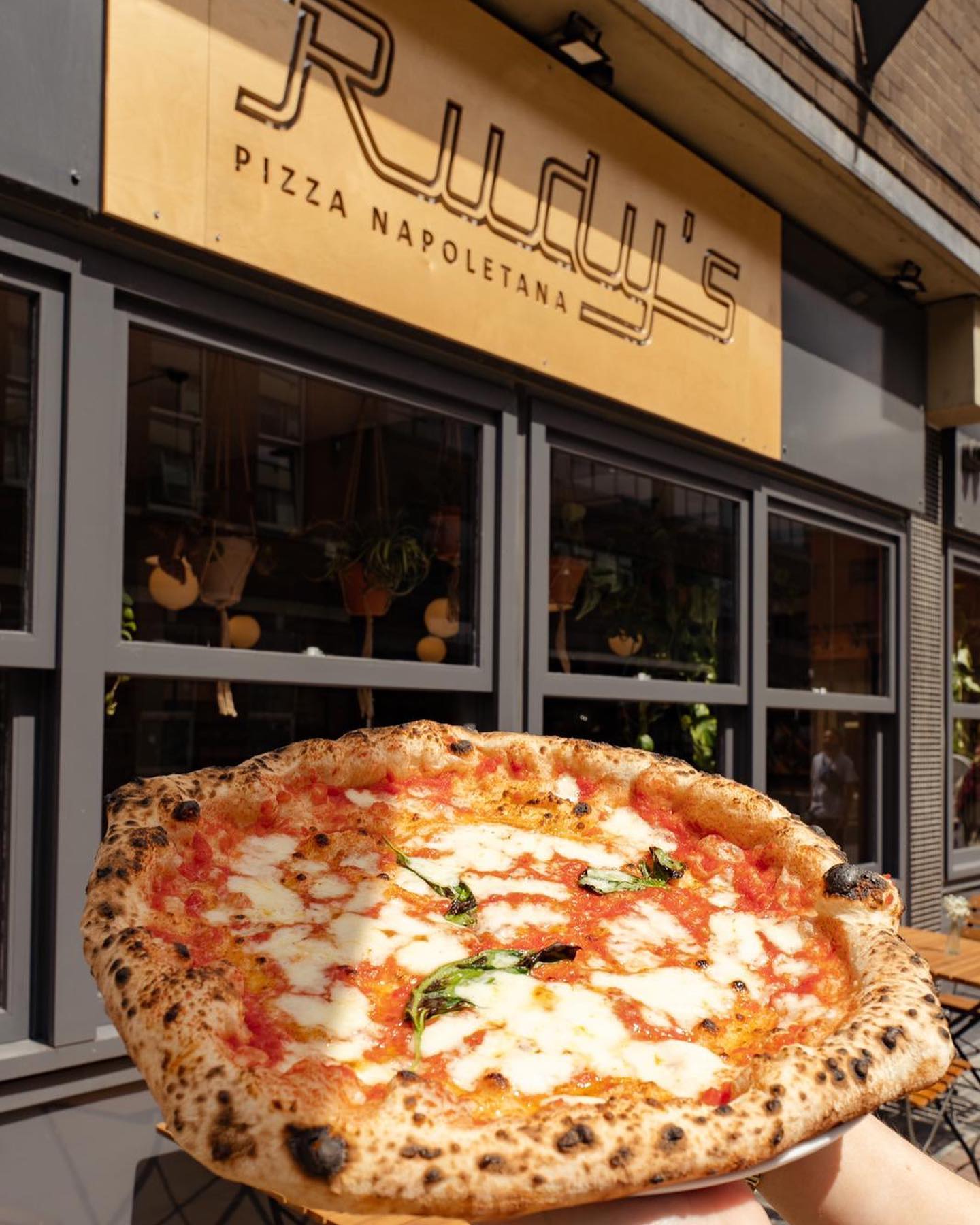 A freshly baked Neapolitan pizza with melted mozzarella, tomato sauce and basil held up in front of a restaurant exterior with a sign reading “Rudy’s Pizza Napoletana.”