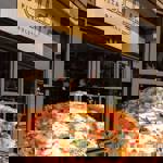 A freshly baked Neapolitan pizza with melted mozzarella, tomato sauce and basil held up in front of a restaurant exterior with a sign reading “Rudy’s Pizza Napoletana.”