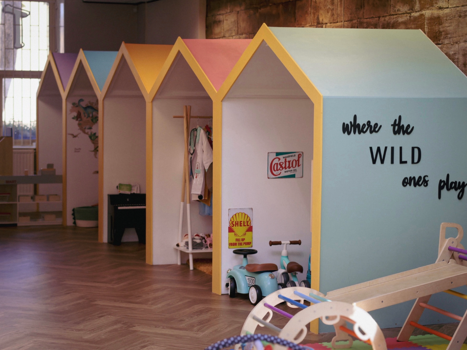 Indoor play area featuring a row of small, house-shaped structures with pastel-coloured roofs and yellow trim. Each section contains toys and play equipment, including a miniature piano, dress-up clothes, and a turquoise ride-on scooter. The front wall of the nearest structure is painted light blue with the phrase “where the WILD ones play” in black lettering. The floor has a herringbone wood pattern, and additional toys are visible in the foreground.
