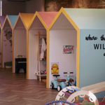 Indoor play area featuring a row of small, house-shaped structures with pastel-coloured roofs and yellow trim. Each section contains toys and play equipment, including a miniature piano, dress-up clothes, and a turquoise ride-on scooter. The front wall of the nearest structure is painted light blue with the phrase “where the WILD ones play” in black lettering. The floor has a herringbone wood pattern, and additional toys are visible in the foreground.