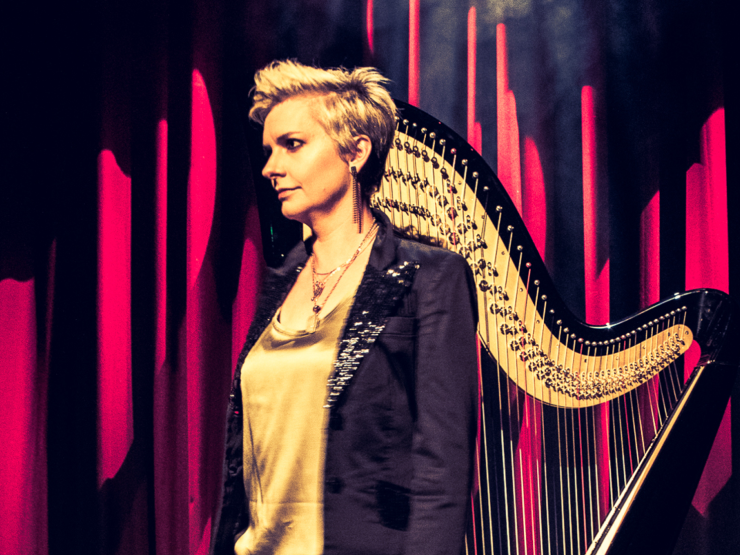 A person stands in front of a large harp on a stage with dramatic red curtains in the background. The harp has a curved black frame with golden strings, and the lighting creates a warm, theatrical atmosphere. The individual is wearing a light top with a dark jacket featuring sequinned details, adding to the performance setting.