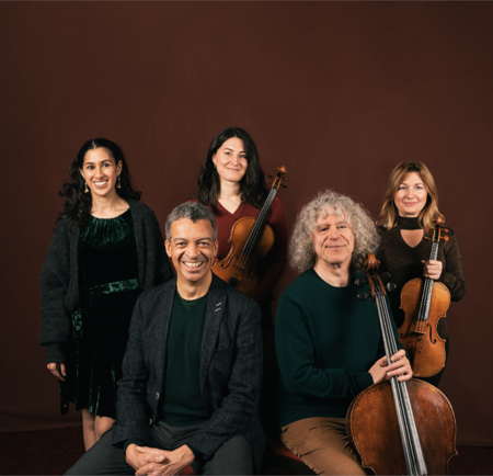 Five people ( 3 women and 2 men) are posing as a group. Two of the women are holding violins and one of the men has a cello. 