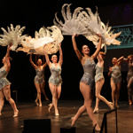 A group of stage performers dance in coordinated formation, each wearing a silver, sparkling costume. They hold large white feathered fans raised above their heads. The stage lighting highlights the movement of the fans, and projection text is visible in the background. Credit: Green Vision Photography
